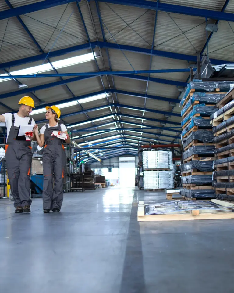 a production hall where a man and a woman wearing yellow helmets are walking and talking